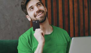 Um homem branco, de cabelos e barba castanho escuro, está usando uma camisa verde e segurando o scale, cartão de crédito com limite flexível da conta simples
